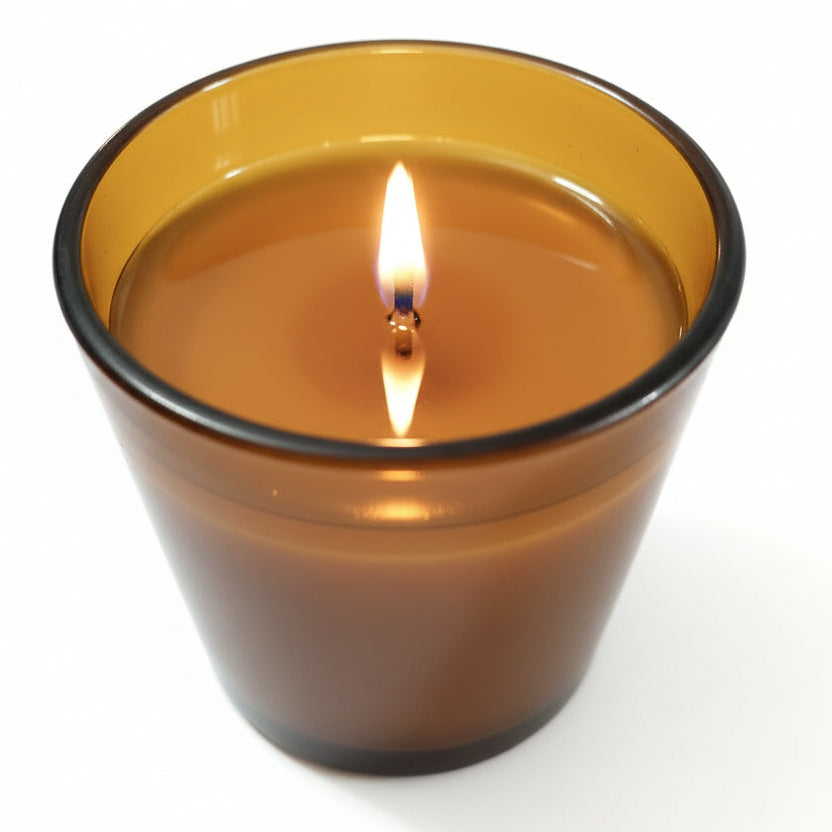 Lit natural wax candle in amber glass container on a white background