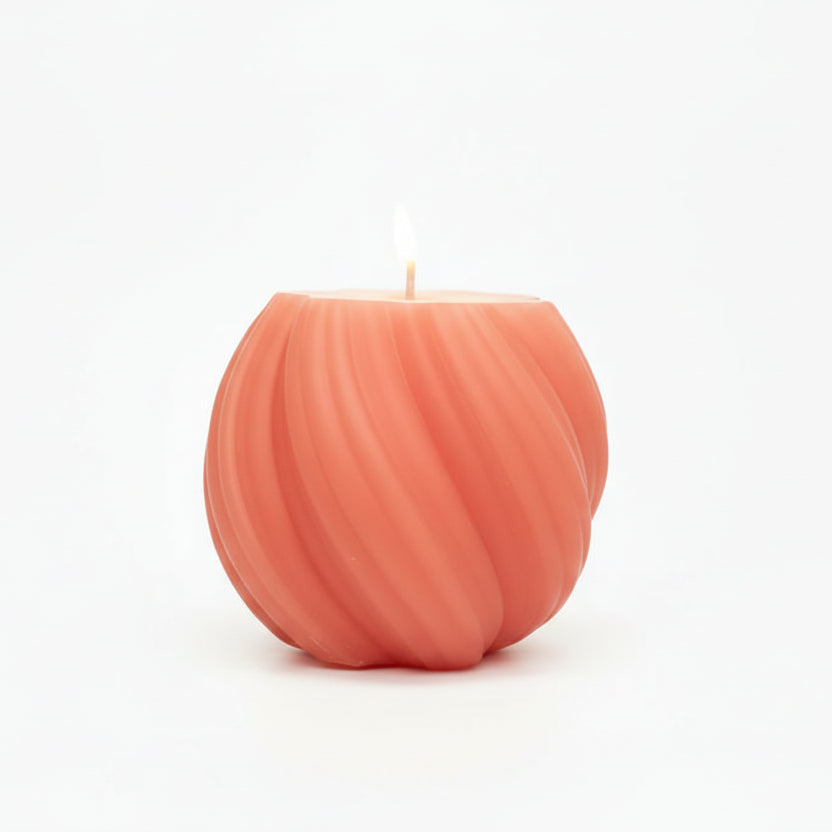 Round sculptural swirl candle in coral color, lit, on white background